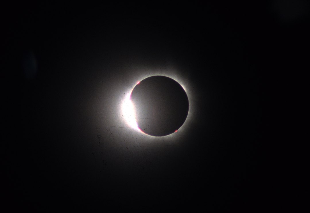 Photographie d'une éclipse totale : un point de lumière très vif au bord gauche du disque lunaire noir, entouré d'un anneau fin et lumineux. Deux petites taches roses sont visibles sur le pourtour.